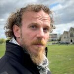 Photo of Bittor, North London SLC member. Bittor is standing in a grassy, open space with buildings and a church visible in the background. He has curly hair and a short beard, and is looking towards the camera, smiling.