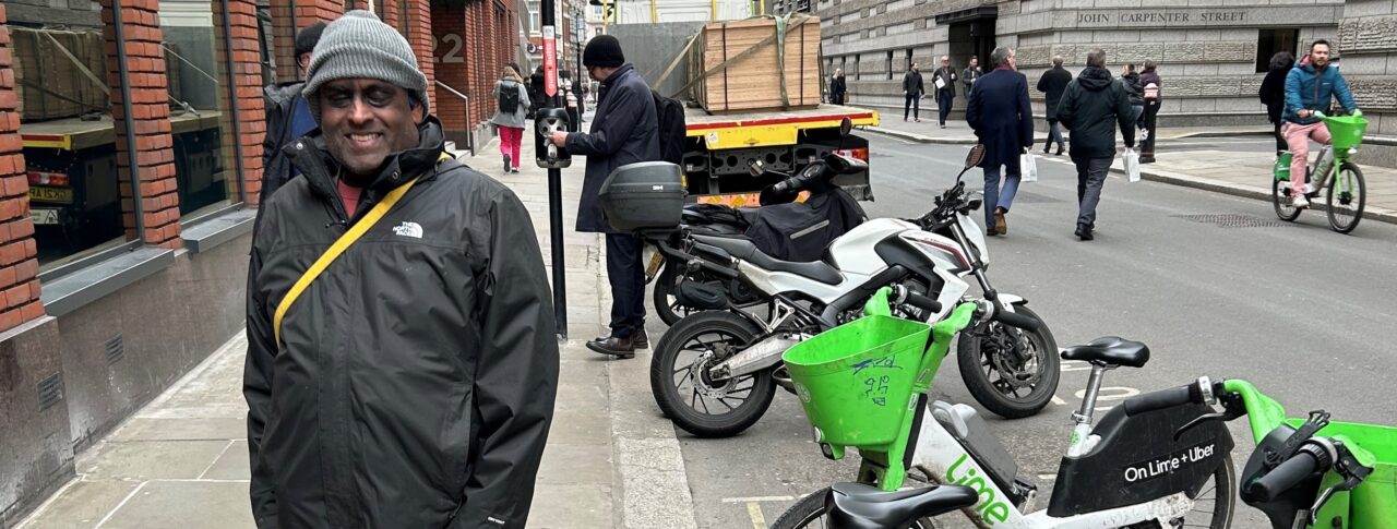 SW London member, Haren, is standing next three Lime bikes with one parked over the curb and protruding onto the path. Haren is with his guide dog, Addy.