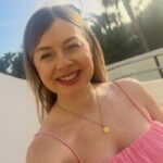 Selfie photo of Katharine, SE London SLC member. She is standing outdoors in a pink, strappy top and a gold necklace. She has long, brown hair. Behind her are palm trees, a white wall, and soft sunlight in the sky.