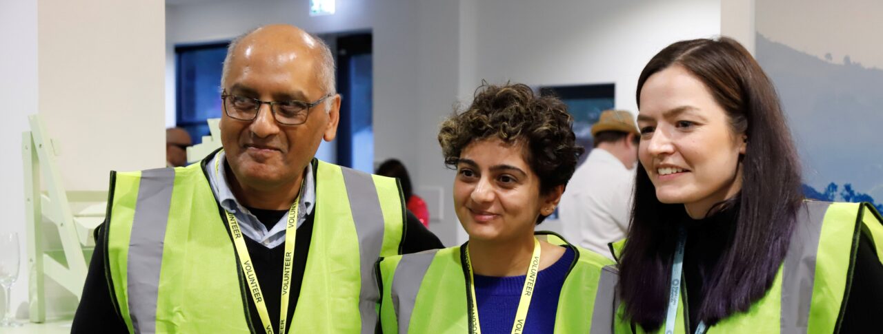 A male and two female sighted guides and stood together at a Sight Loss Council conference. They are wearing yellow tabards and lanyards. They are stood in a line looking to their right, and have happy expressions on their faces.