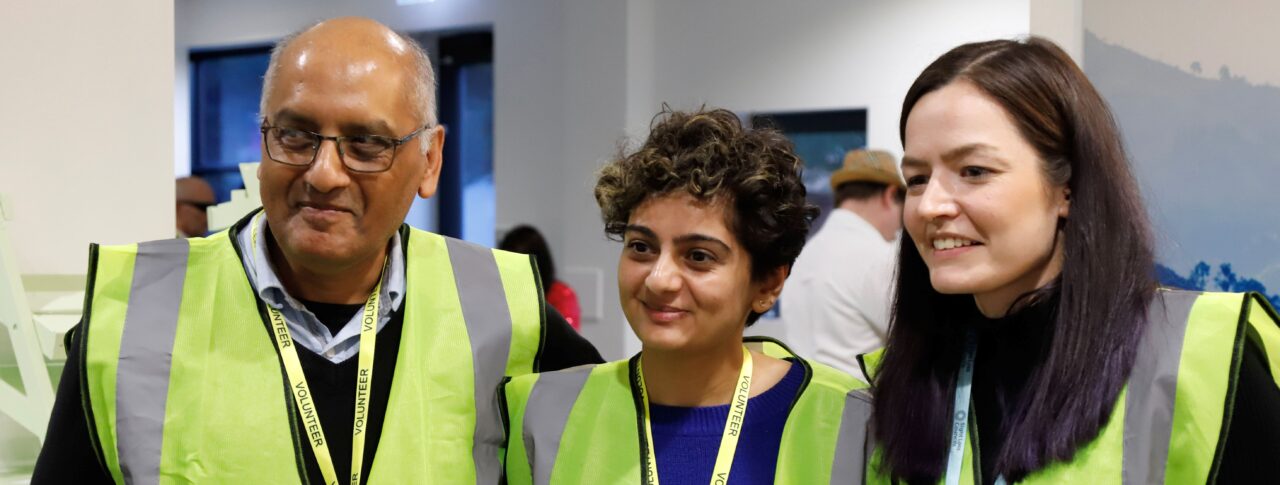 A male and two female sighted guides and stood together at a Sight Loss Council conference. They are wearing yellow tabards and lanyards. They are stood in a line looking to their right, and have happy expressions on their faces.