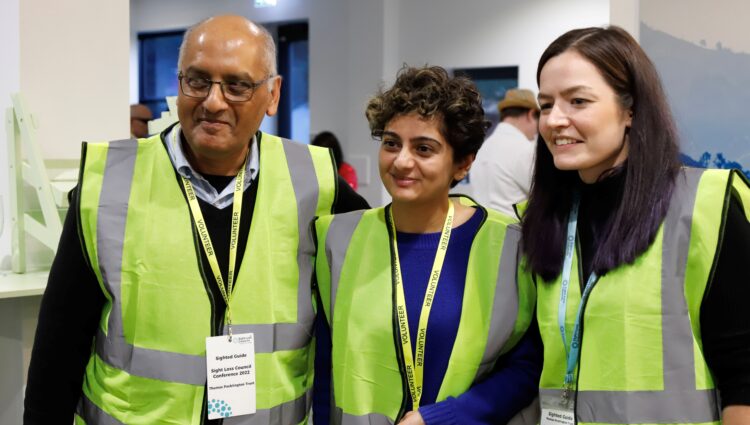 A male and two female sighted guides and stood together at a Sight Loss Council conference. They are wearing yellow tabards and lanyards. They are stood in a line looking to their right, and have happy expressions on their faces.