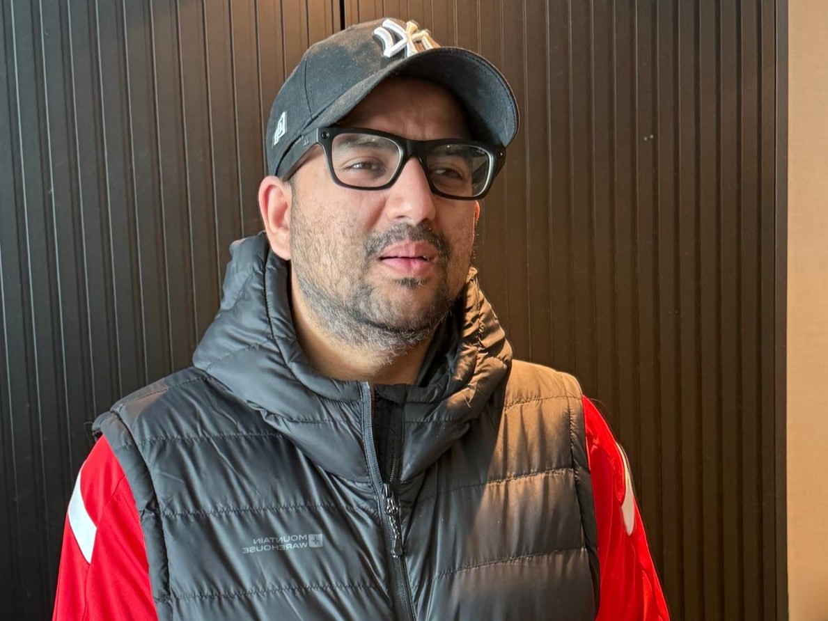 Abu-bakr, Greater Manchester SLC member, stands in front of a dark wall. He is wearing a black baseball cap, dark framed glasses, and padded gilet. His head is slightly turned to the side.