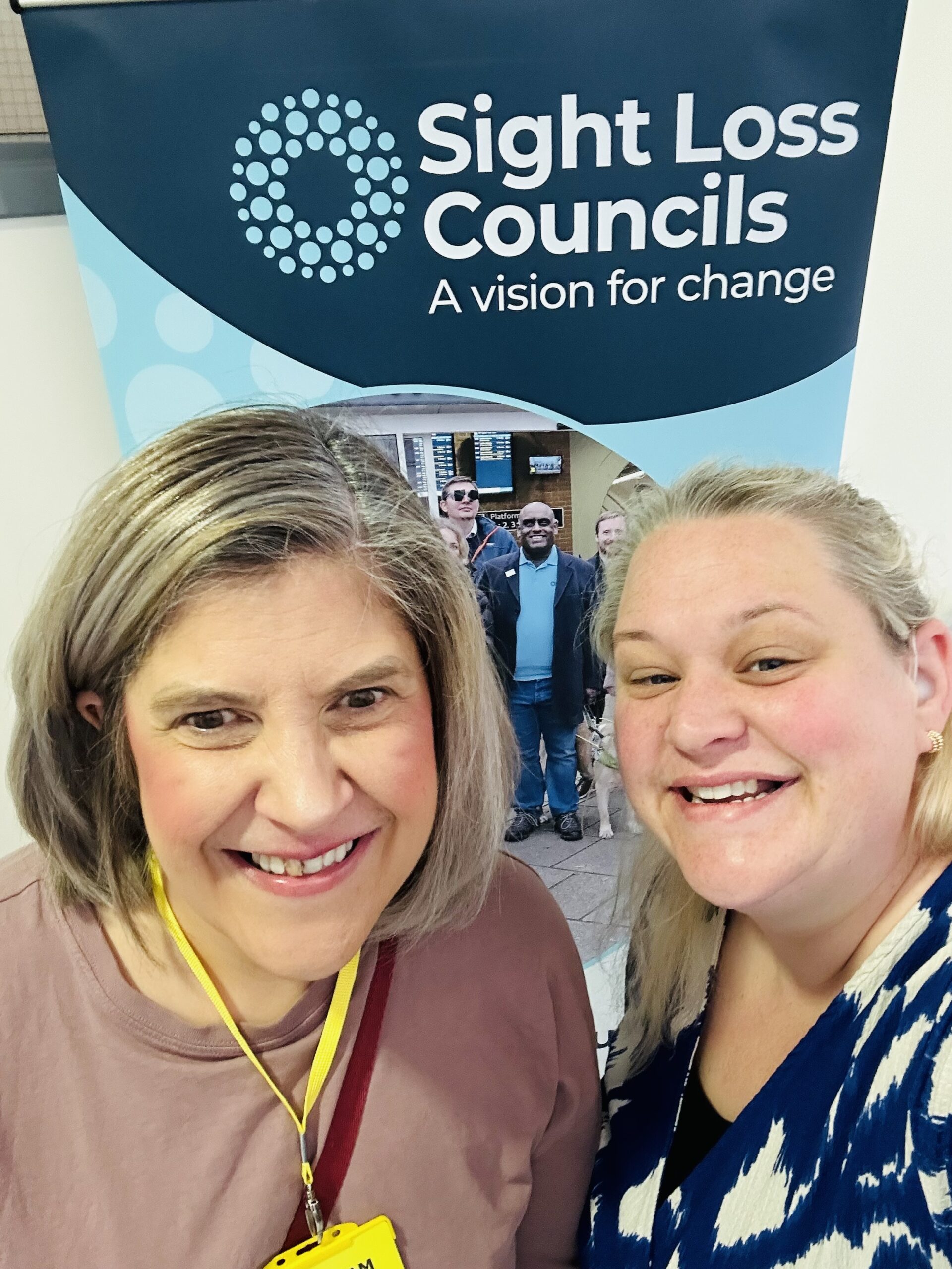 Selfie of Helen and Dannie, Derbyshire SLC members, stood in front of the Sight Loss Councils banner.