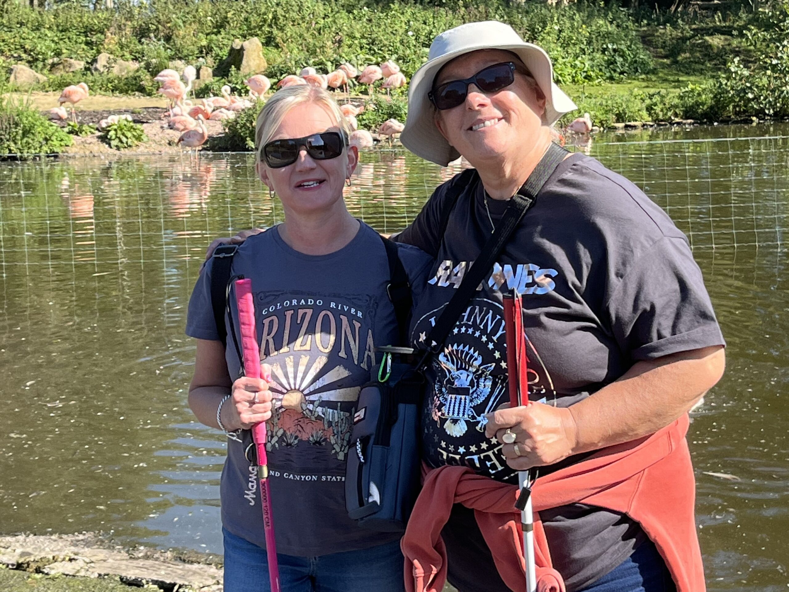 Kelly Barton, Engagement Manager for North West England, stands with Dawn from Lancashire SLC in front of a large pond. Both wear sunglasses, hold their long canes, and are smiling at the camera.