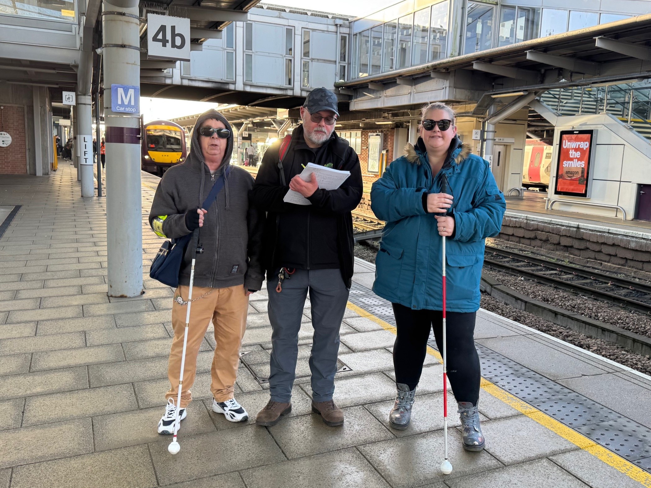 Dale and Dannie, with support worker John, stand on a platform at a railway station. Dale and Dannie each hold a long cane and are smiling at the camera. John is looking down, making notes on an A4 pad.