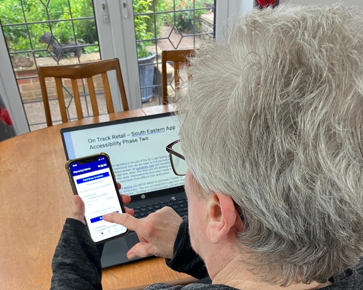 Leslie, North Yorkshire SLC member, is pictured at her table, holding a smartphone. The smartphone displays a rail ticket booking app with a prominent “Find times & tickets” button. In front of the person is an open laptop showing a document titled “On Track Retail – South Eastern App Accessibility Phase Two.”