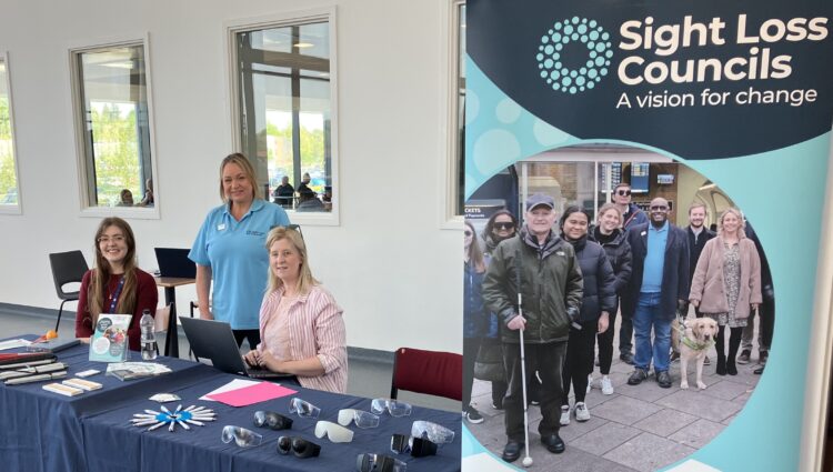 Photo taken at Royal Stoke Hospital during Patient Experience Week. From left to right: Madeleine Roche, Patient and Public Involvement and Engagement Co-ordinator is seated behind an information table, PJ, SLC Coordinator for West Midlands is standing behind the table, and Louise Connop, Senior Engagement Manager for South England, is seated beside a laptop. A SLC banner stands to the right of the group and the table displays information materials and simulation glasses.