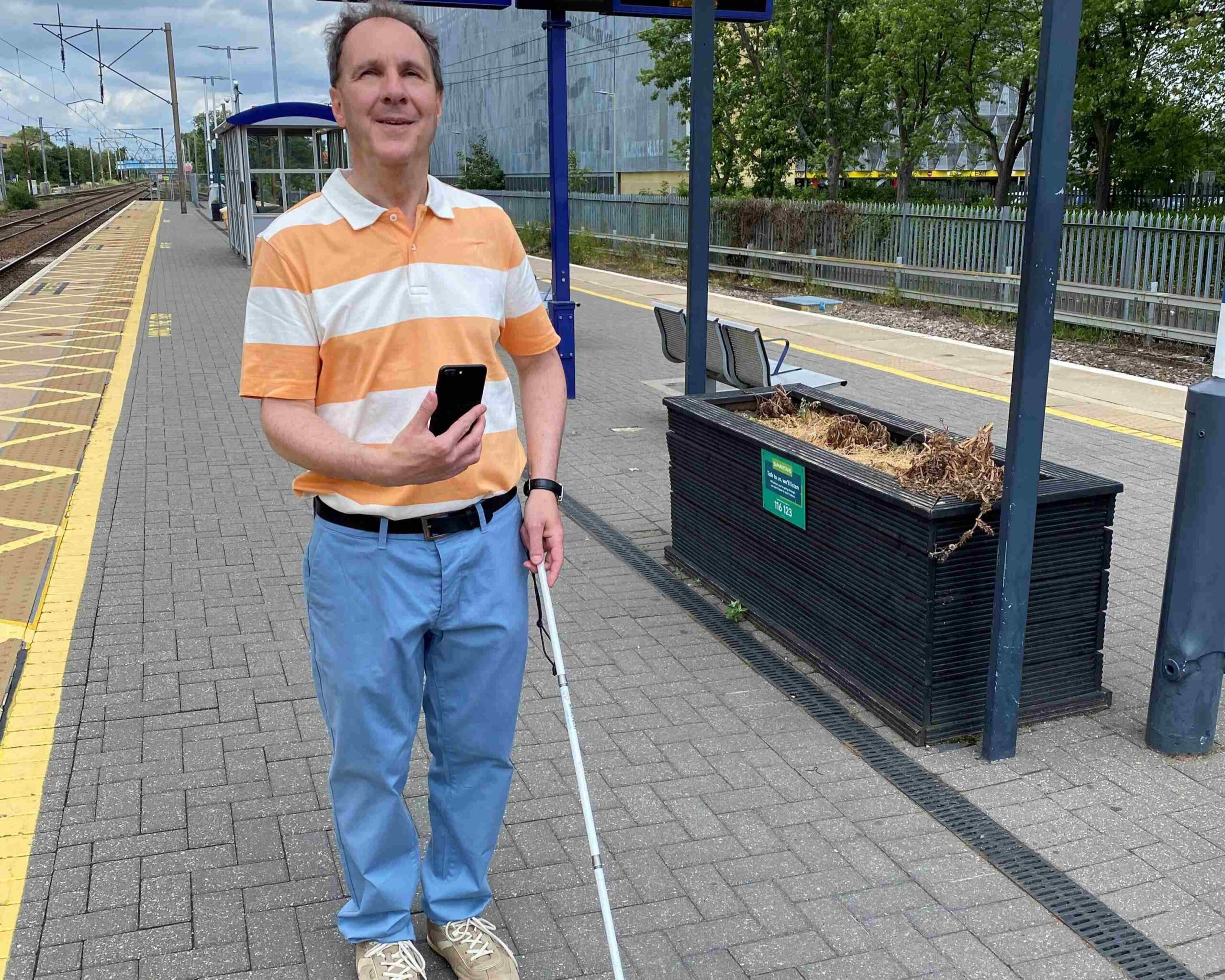 Paul, Bedfordshire and Hertfordshire SLC member, is standing on a platform at Stevenage train station during the AIRA app testing. He is holding his long cane in one hand, and his phone up in the other.