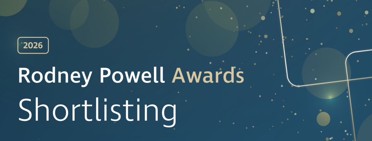 Graphic banner with a dark blue gradient background featuring soft, translucent circular shapes and scattered gold dots. On the left, a gold‑outlined rounded rectangle contains the text “2026.” Large white and gold text reads “Rodney Powell Awards,” with “Shortlisting” displayed prominently below in white. Decorative curved gold lines sweep across the right side, with a subtle glowing effect near the lower right corner, adding a celebratory, polished feel.
