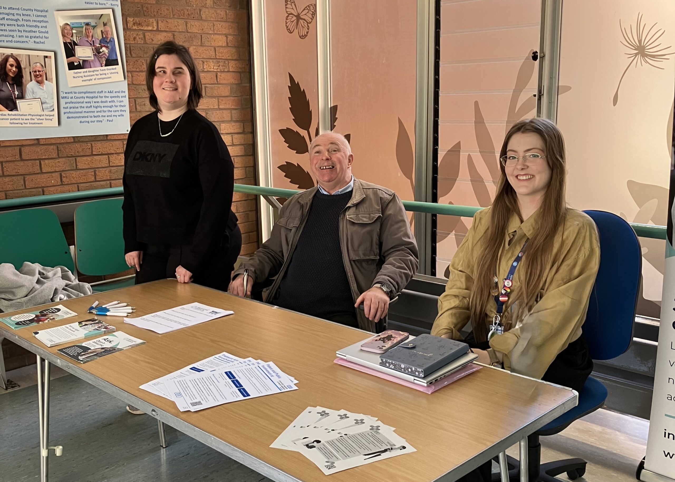 Photo taken at University Hospital North Midlands during Patient Experience Week. From left to right: Klaudia and Geoff, Staffordshire SLC members, and Madeleine Roche, Patient and Public Involvement and Engagement Co-ordinator. Geoff and Madeleine are seated at a table while Klaudia stands beside them. All three are smiling at the camera.
