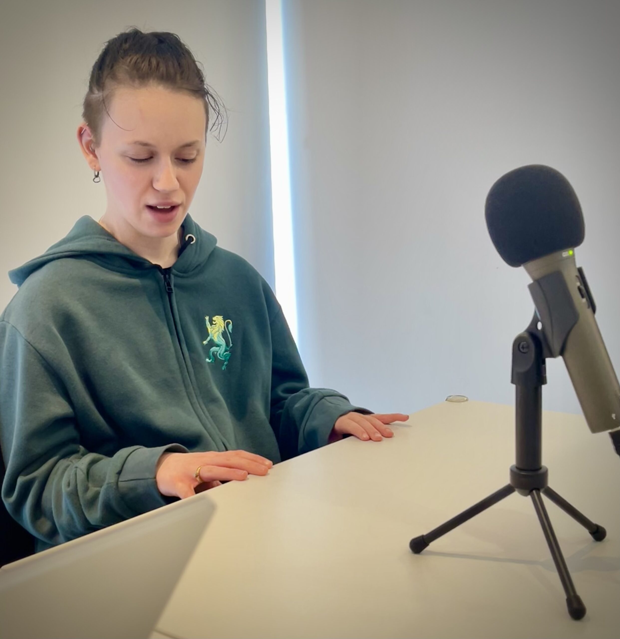Faith, Student Voices member, sits at a wooden desk with a microphone on a stand.
