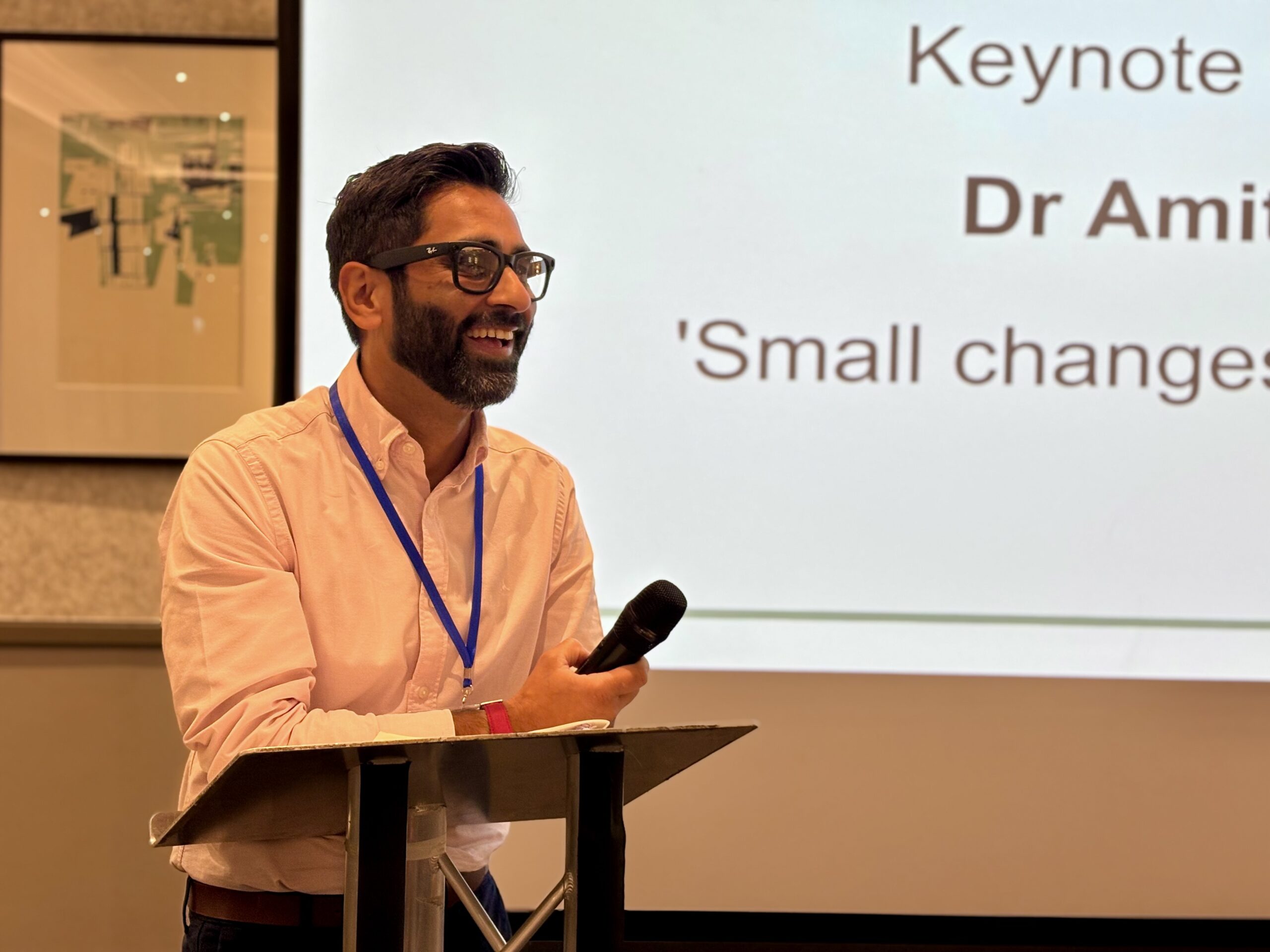 Dr Amit Patel, also known as Blind Dad, stands at a lectern in the main conference room during a keynote speech. Wearing glasses, a light pink shirt and a conference lanyard, he holds a handheld microphone and leans forward as he speaks. A large projection screen behind him displays partially visible presentation text, including the word “Keynote”, while framed artwork is visible on the wall to one side.