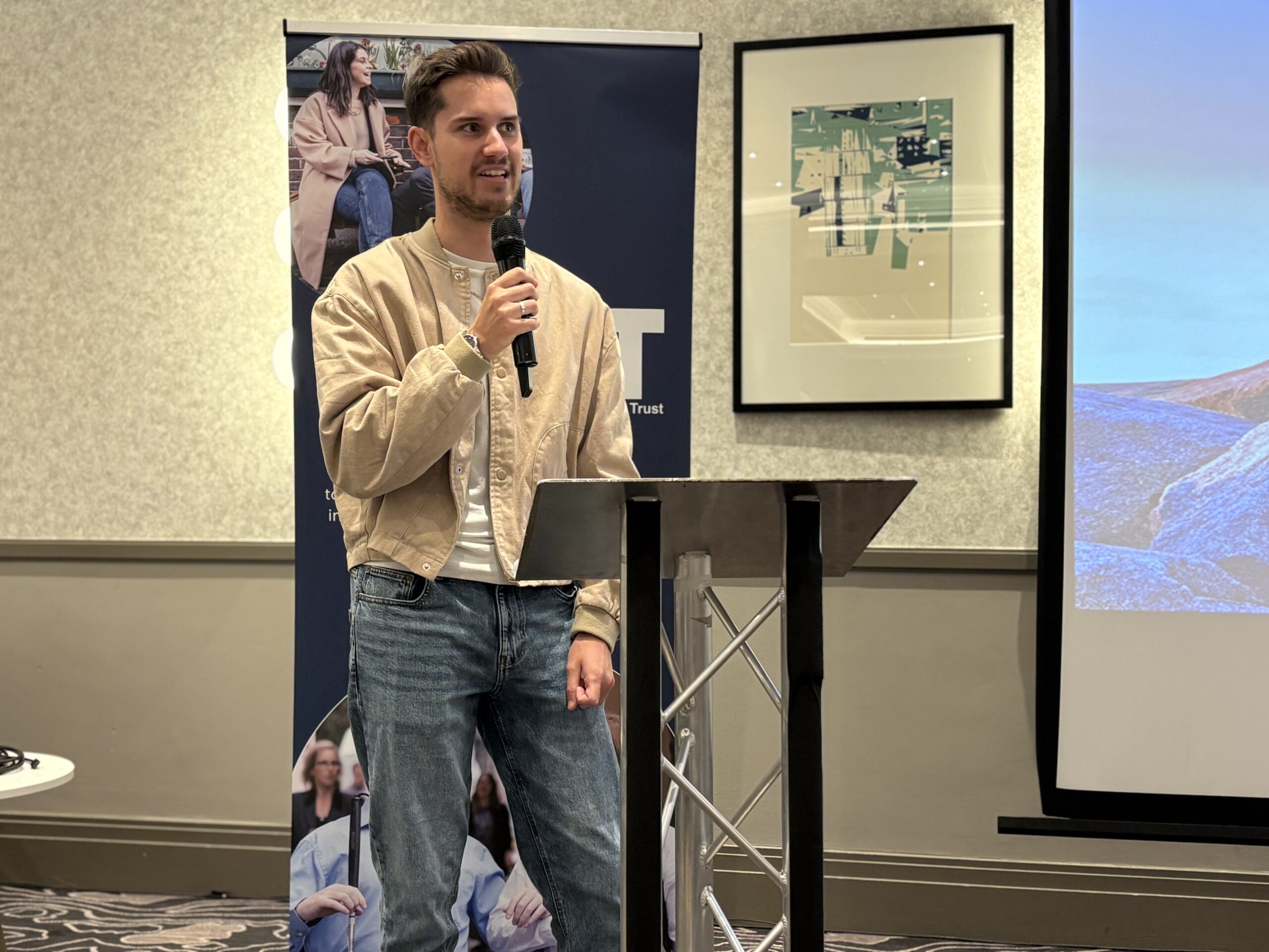 Close up photo of Toby Addison during this keynote speech during the closing session of the 2026 SLC Conference. He is stood in front of a TPT banner, speaking into a microphone. He is wearing a stone coloured jacket.