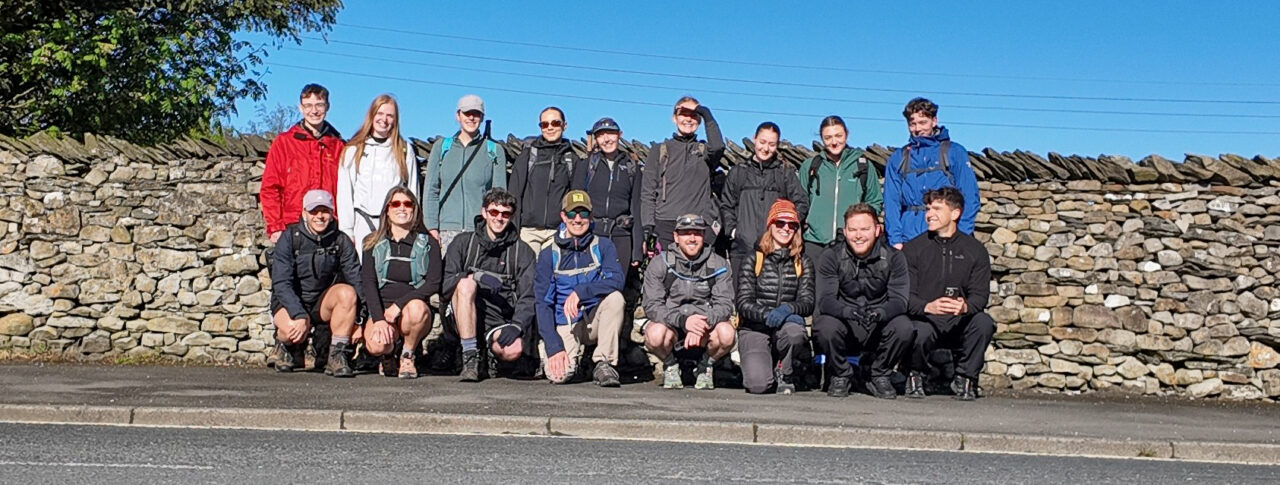 17 staff members from GT3 Architects stand beside a dry stone wall on a sunny day. Some are standing and others are crouching at the front. They wear outdoor clothing, backpacks and walking gear. The group is standing on a pavement next to a road, with trees, a stone wall and a clear blue sky in the background.
