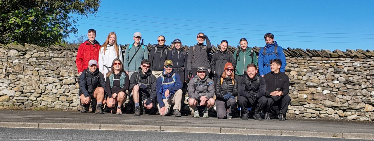 17 staff members from GT3 Architects stand beside a dry stone wall on a sunny day. Some are standing and others are crouching at the front. They wear outdoor clothing, backpacks and walking gear. The group is standing on a pavement next to a road, with trees, a stone wall and a clear blue sky in the background.