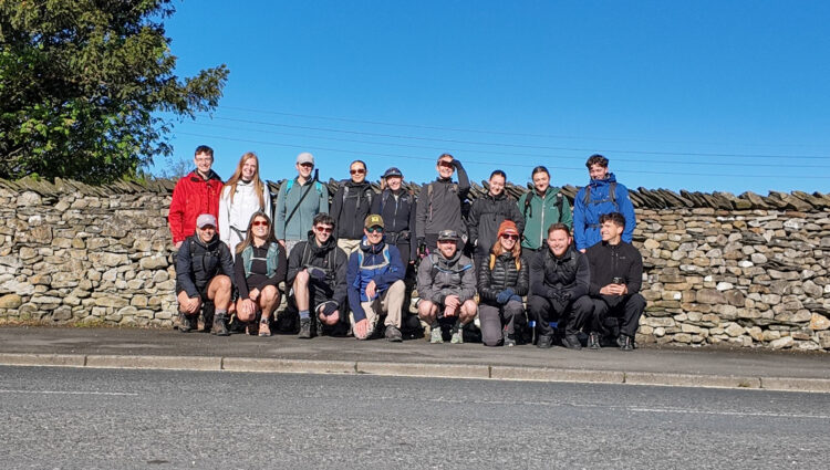 17 staff members from GT3 Architects stand beside a dry stone wall on a sunny day. Some are standing and others are crouching at the front. They wear outdoor clothing, backpacks and walking gear. The group is standing on a pavement next to a road, with trees, a stone wall and a clear blue sky in the background.