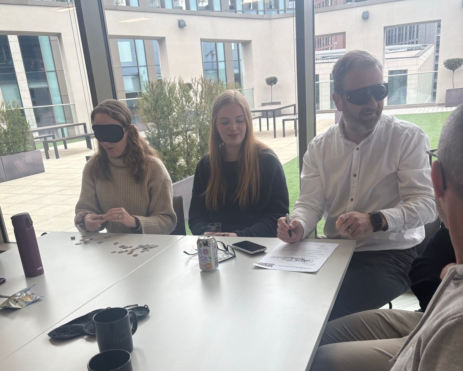 Three staff members sit around a square table during a vision awareness session. One is wearing a blind fold and doing a task with coins. Another is wearing simulation glasses, trying to complete a paper form.
