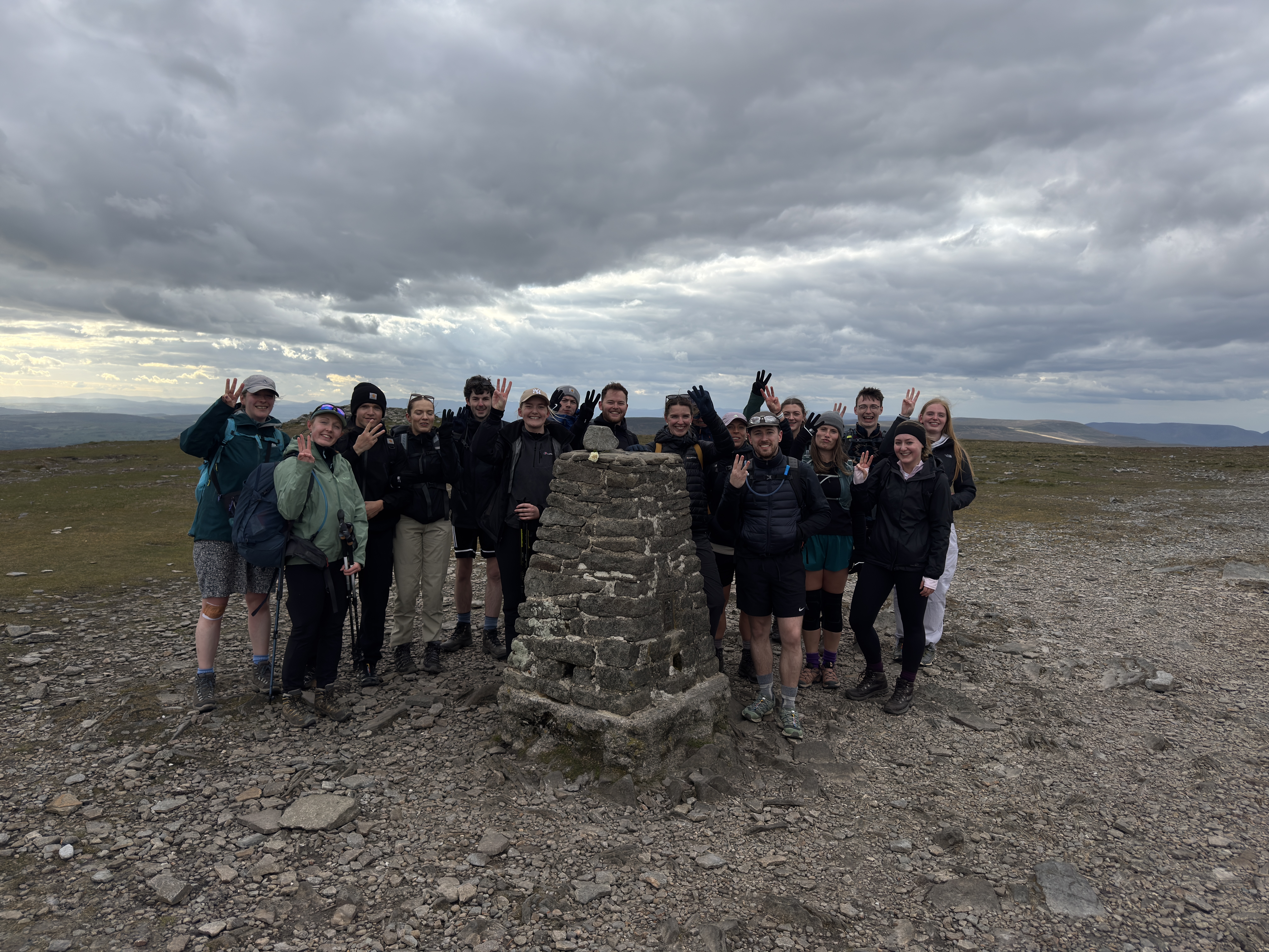 Staff members from GT£ Architects pose around a stone summit marker on a rocky hilltop. Several people raise four fingers towards the camera. The group is dressed in outdoor clothing and walking gear, with an expansive landscape stretching into the distance beneath dramatic dark clouds.