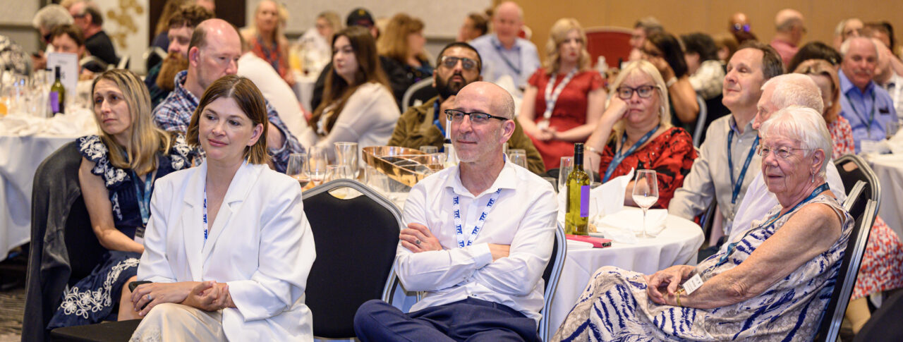 Audience members seated around round tables at the Rodney Powell Awards event. In the foreground, three attendees sit facing the front of the room, while additional guests fill the background. Tables are set with glasses, bottles and table numbers in a hotel conference venue with patterned carpet and warm lighting. The image captures the audience's attention during a speech.