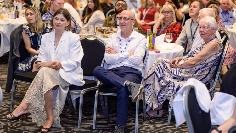 Audience members seated around round tables at the Rodney Powell Awards event. In the foreground, three attendees sit facing the front of the room, while additional guests fill the background. Tables are set with glasses, bottles and table numbers in a hotel conference venue with patterned carpet and warm lighting. The image captures the audience's attention during a speech.