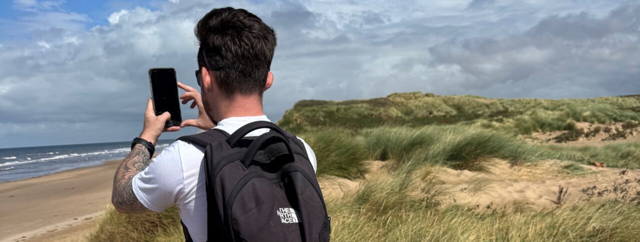 David Parfitt, SLC Coordinator for North West England, stands with a backpack on his back, on sand dunes at Formby Beach. He holds up a smartphone to take a photo of the sea. Grassy dunes line the right side of the image.