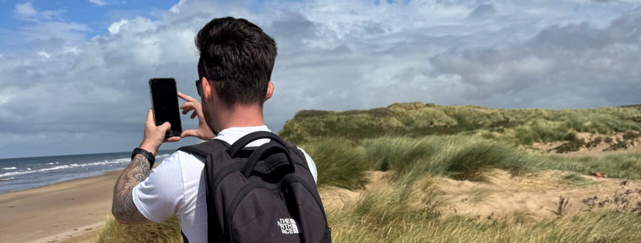 David Parfitt, SLC Coordinator for North West England, stands with a backpack on his back, on sand dunes at Formby Beach. He holds up a smartphone to take a photo of the sea. Grassy dunes line the right side of the image.