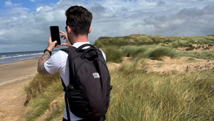 David Parfitt, SLC Coordinator for North West England, stands with a backpack on his back, on sand dunes at Formby Beach. He holds up a smartphone to take a photo of the sea. Grassy dunes line the right side of the image.