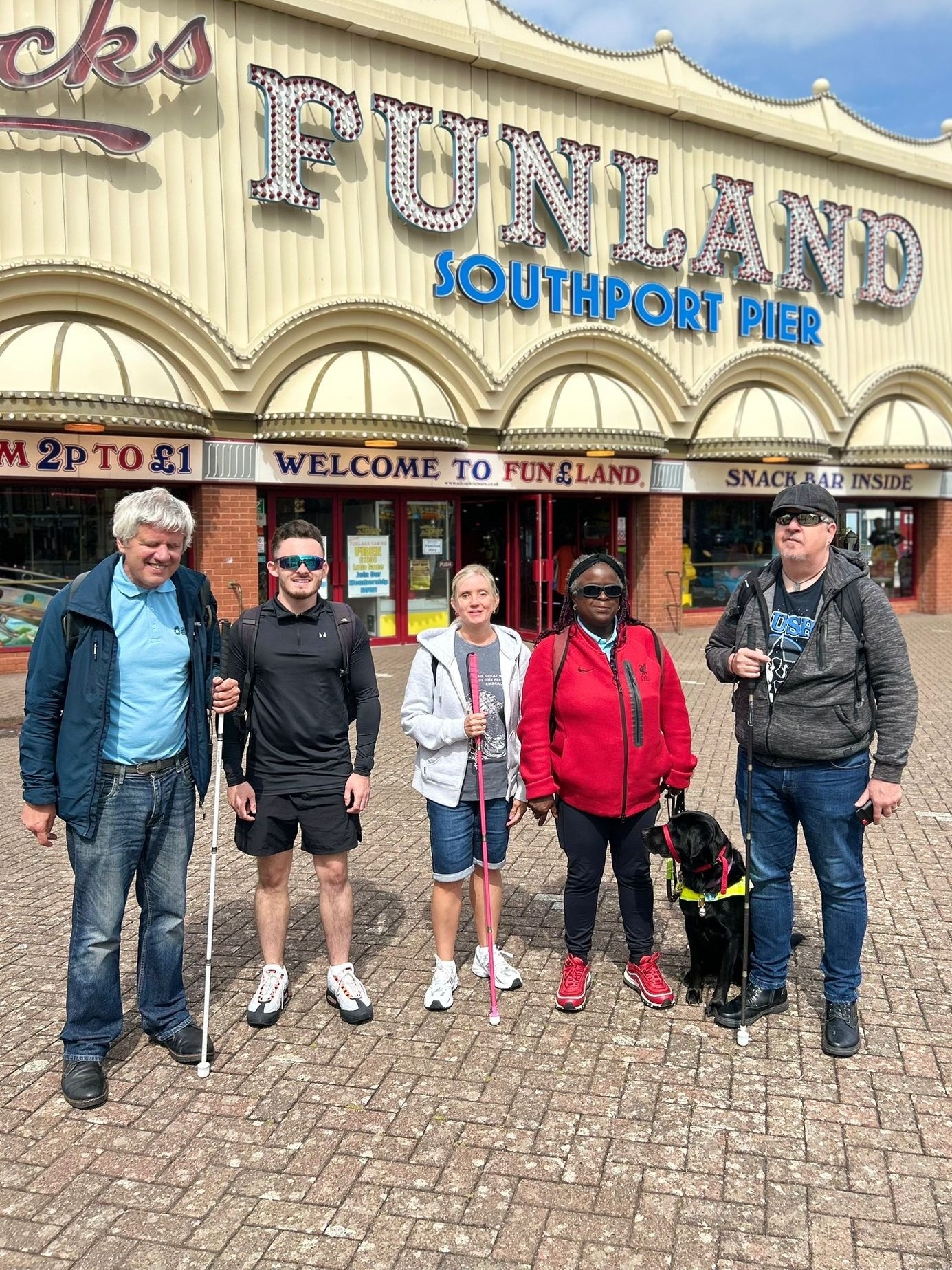 Merseyside SLC members stand together underneath the Funland Southport Pier sign. From left to right: Mick, David Parfitt, SLC Coordinator for North West England, Kelly Barton, Engagement Manager for North West England, Naomi and Guide Dog Dottie, and James. Mike, Kelly, and James each hold a long cane in their hands.