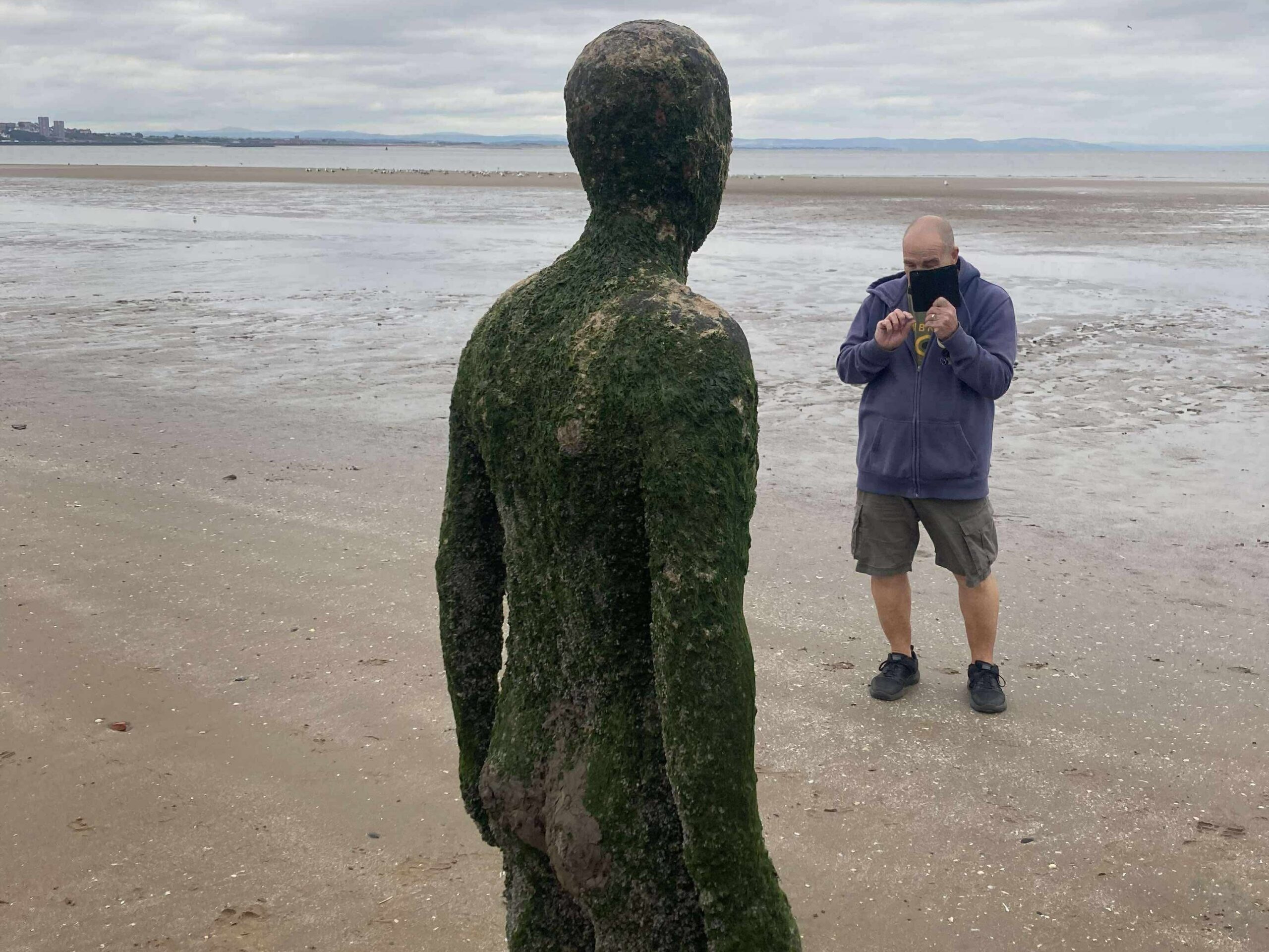 Mick, Merseyside SLC member, stands on the sand at Crosby Beach, taking a photo of an Andrew Gormley, cast iron statue, which faces the sea. Mick is looking down towards his phone, the tide is out and seen in the distance.