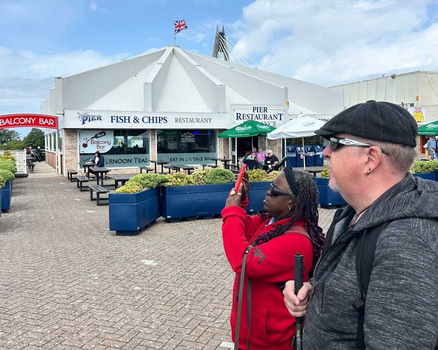 Merseyside SLC members, Naomi and James, during the Through Our Eyes project. They stand outside a Southport seafront restaurant, with Naomi taking a photo on a smartphone. James stands next to her, looking on, and holds a long cane.