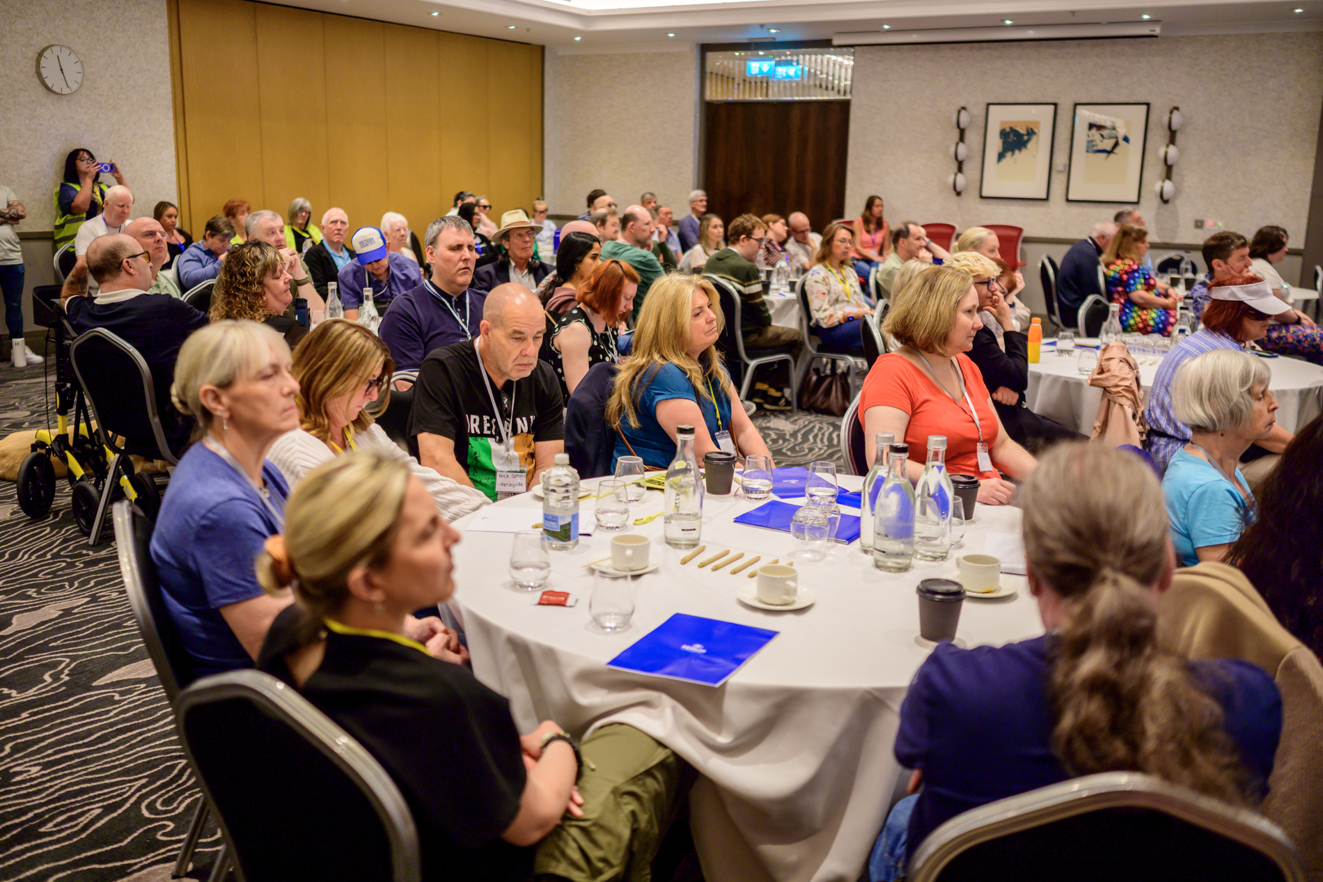 A large group of conference attendees are seated around round tables in the main conference room during the opening address. Water bottles, glasses, notebooks and conference materials are arranged on the tables. A mobility scooter is visible on the left, and a person at the back of the room is taking a photograph. Framed artwork and wall lights are visible on the walls behind the audience.