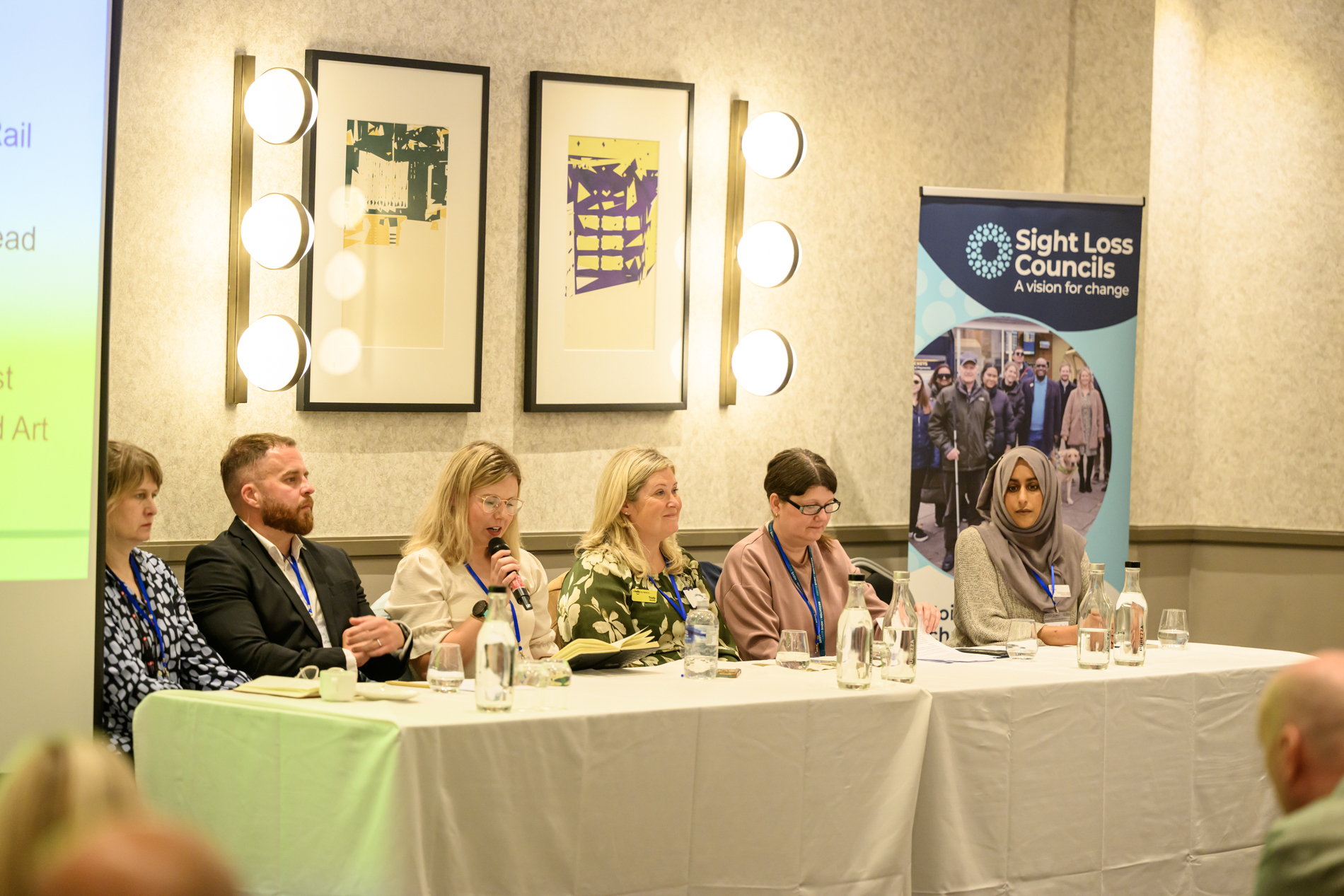 A panel of six speakers seated behind a white-clothed table during the Storytellers Panel. Trudy Taylor speaks into a handheld microphone while the other panellists listen. Water bottles, glasses and notes are arranged on the table. A Sight Loss Councils banner stands behind the panel, and part of a presentation screen is visible on the left. From left to right are Tom Sunley, Lauren Lett, Trudy Taylor, Victoria Garcia and Shahiesta Raja. Two framed abstract artworks and circular wall lights are displayed on the wall behind the pane