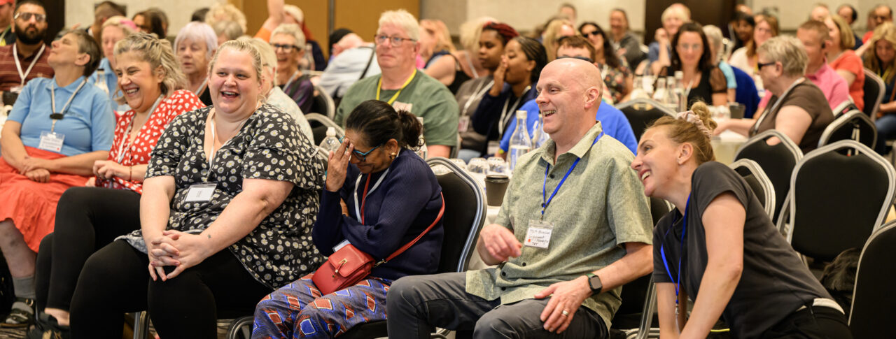 Staff and volunteers seated at the front of a conference room during the 2026 Sight Loss Councils Conference in Leeds, listening to a speaker and laughing as they listen. Delegates gathered around tables in the background.
