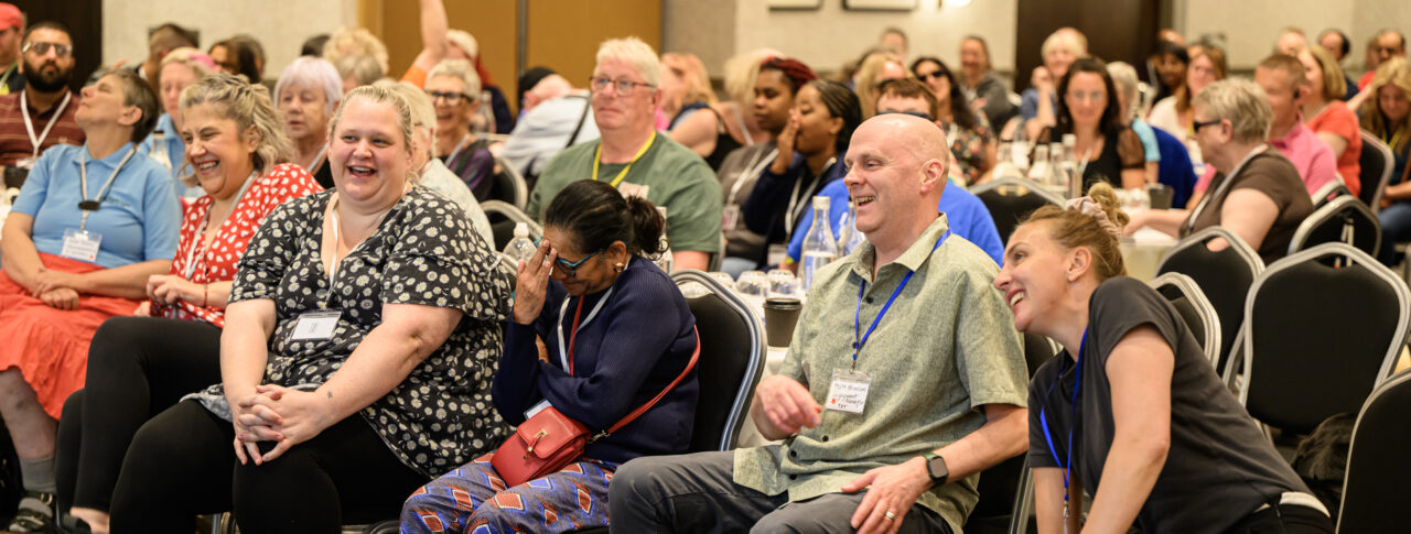 Staff and volunteers seated at the front of a conference room during the 2026 Sight Loss Councils Conference in Leeds, listening to a speaker and laughing as they listen. Delegates gathered around tables in the background.