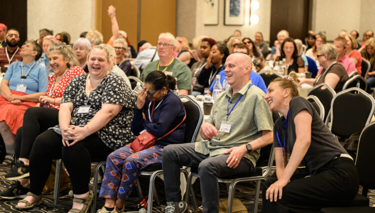 Staff and volunteers seated at the front of a conference room during the 2026 Sight Loss Councils Conference in Leeds, listening to a speaker and laughing as they listen. Delegates gathered around tables in the background.