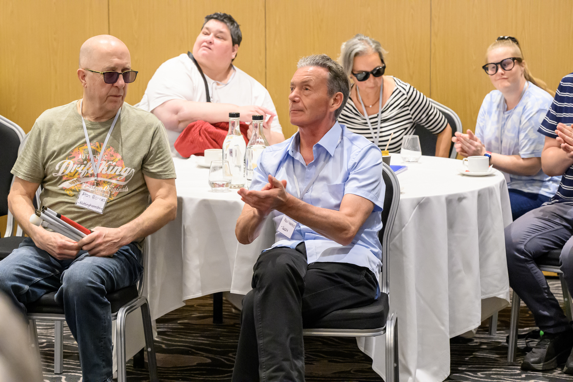 Several conference attendees are seated around round tables in the main conference room. Two attendees sit in the foreground, one holding a folded white cane and the other applauding. Water bottles, glasses and cups are arranged on the tables, and several attendees wearing sunglasses are visible in the background