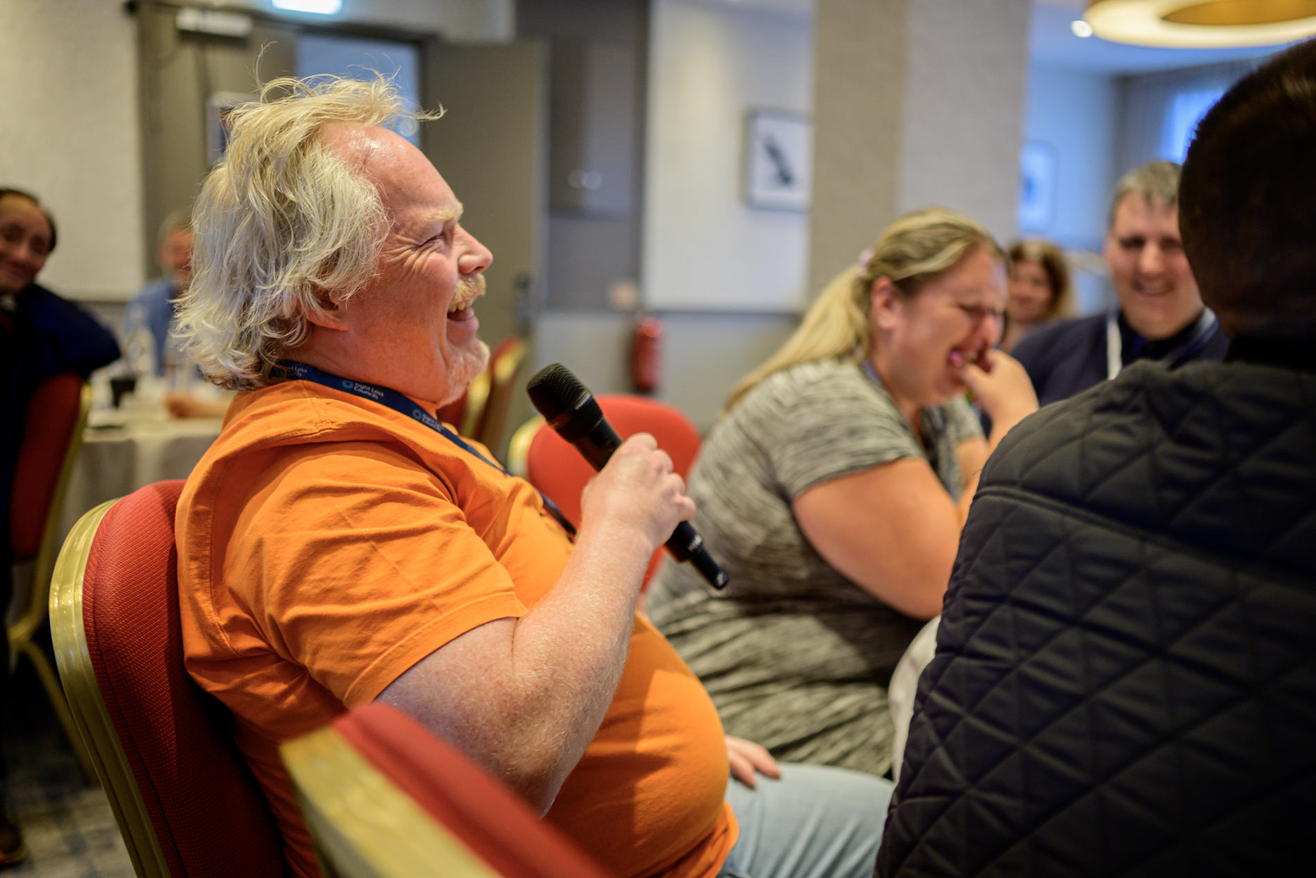 Steve Keith, Birmingham and Black Country SLC member speaks into a handheld microphone during a session at conference. Several other attendees are seated nearby, with tables and chairs visible in the meeting room. The image is taken from the side, with the speaker in the foreground and other participants in softer focus in the background.