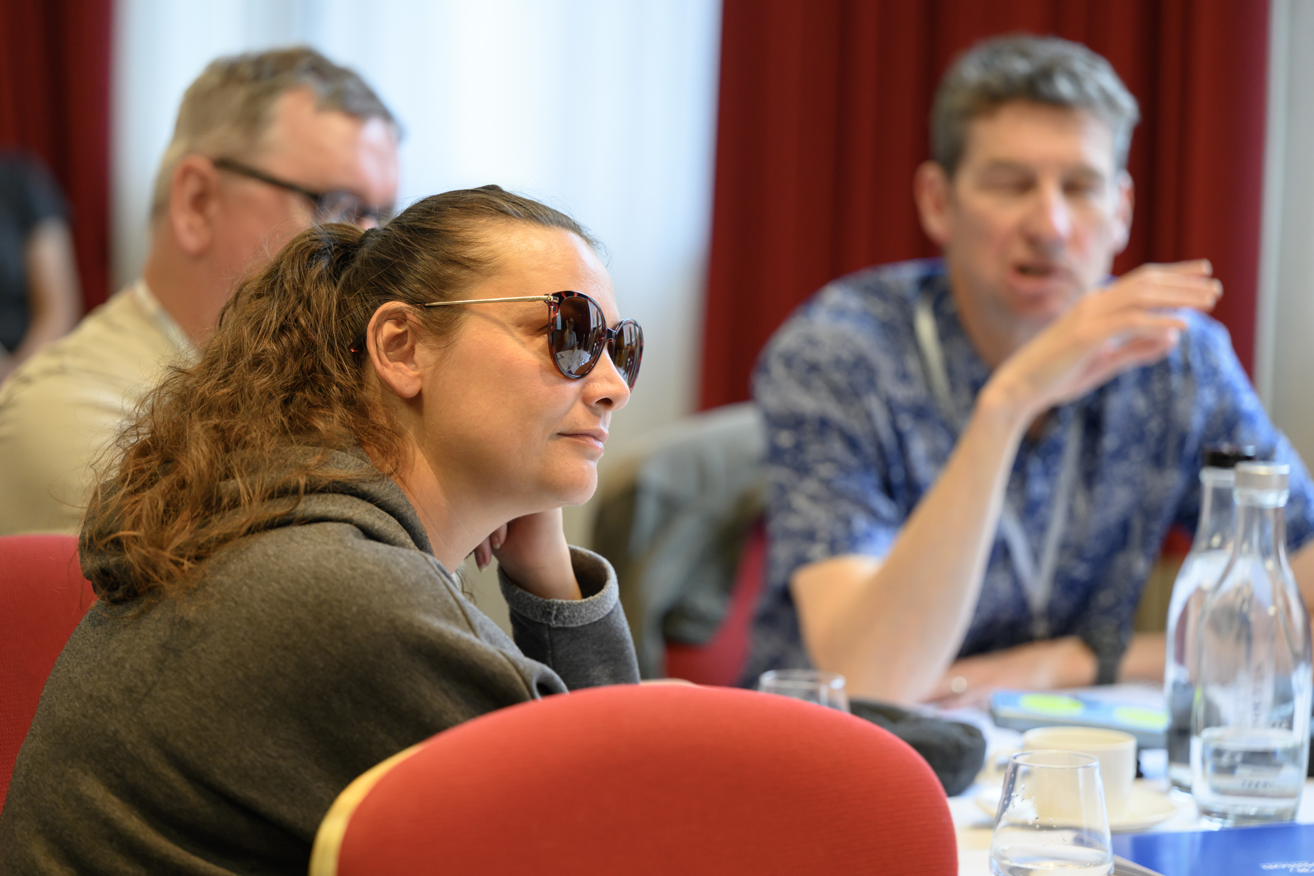 A conference attendee wearing sunglasses sits at a table during a group discussion. Other attendees are seated nearby, slightly out of focus. Water glasses, a bottle and meeting materials are arranged on the table, with red curtains visible in the background.