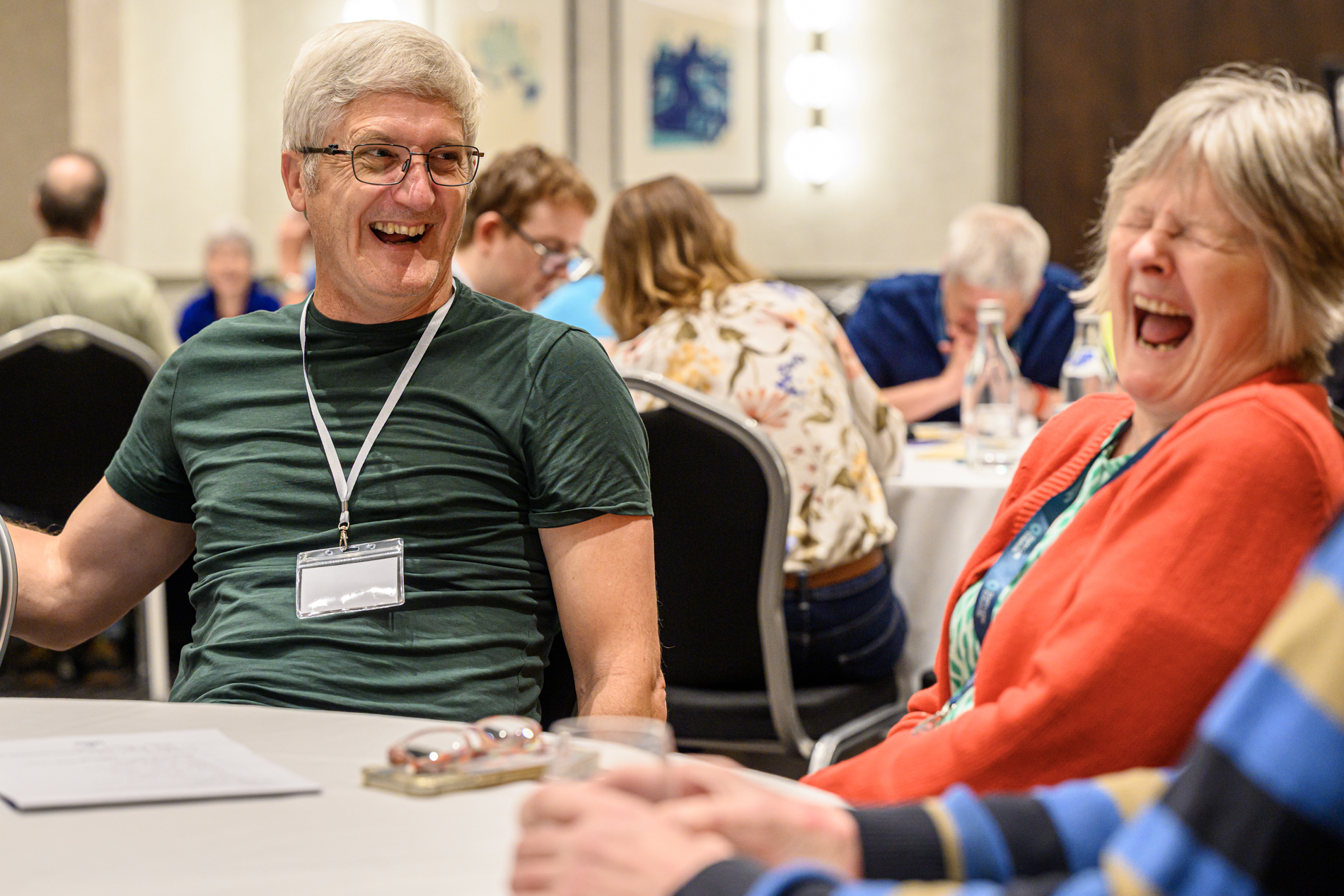Two SLC delegates sit at a table, facing one another in conversation - both are laughing. Other attendees are seated at tables in the background.