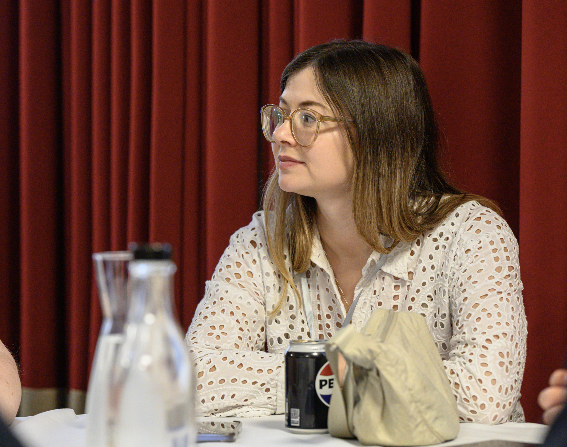 Katharine, SE London SLC member, wears glasses and a white patterned blouse and sits at a table, listening during a discussion. A water bottle, mobile phone, soft drink can and bag are placed on the table in the foreground. Red curtains form the background.