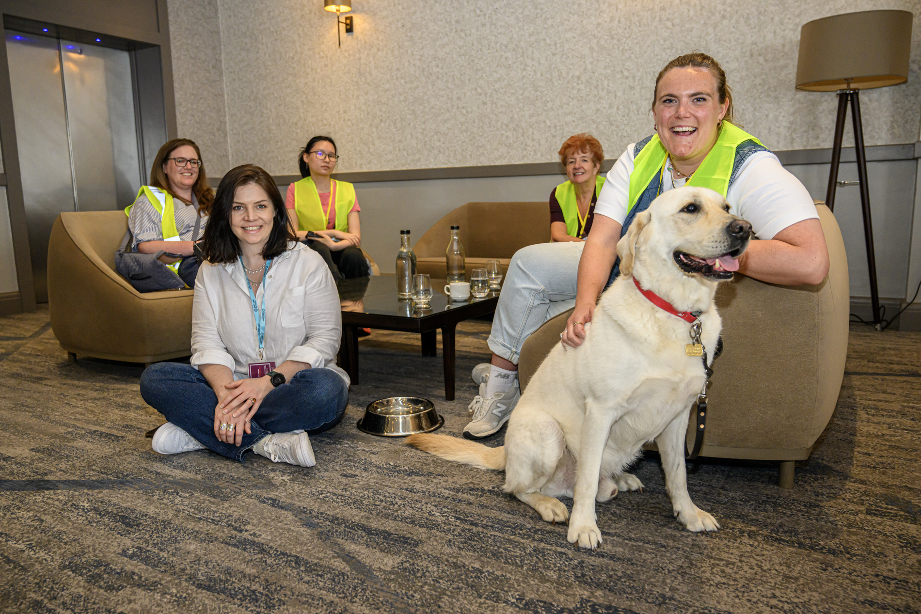A group photo in the reception area of the Hilton Leeds shows Tina sitting cross-legged on the floor in the foreground. Behind Tina, four sighted guides wearing high-visibility yellow vests are seated on sofas and chairs around a low table with water bottles and glasses. A guide dog sits at the front of the image beside a person seated in an armchair.