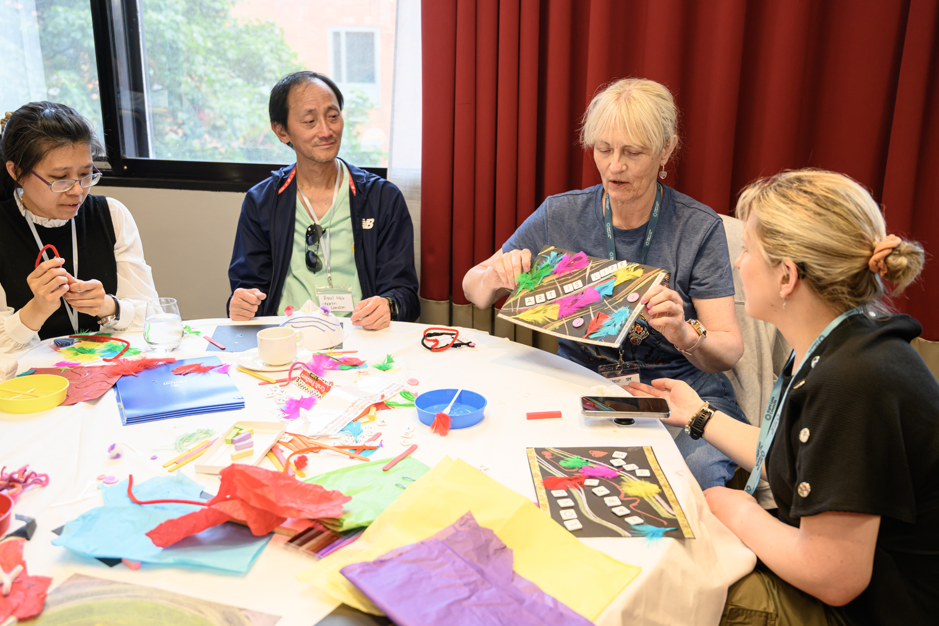 Ching, SE London, Paul, North London, Carolyn, Merseyside SLC, and a sighted companion sit around a table taking part in the What Culture Means to Me workshop. Colourful craft materials, including feathers, tissue paper, pipe cleaners, coloured sticks and paper, are spread across the table. Carolyn holders up her decorated artwork which features bright feathers and the words: "Art is life", whilst others look on.