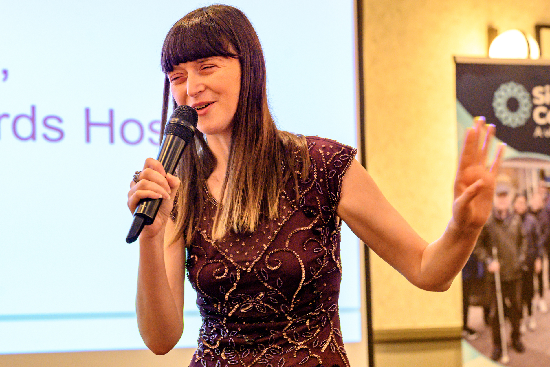 Fern Lulham, host of the 2026 Rodney Powell Awards, holds a handheld microphone as she speaks to the audience. She is wearing a dark patterned sleeveless dress, and gesturing with one hand while addressing the room. Behind her, is a large presentation screen and to the right, a freestanding banner for Sight Loss Councils.