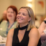 Stevie, South Yorkshire SLC member, at the 2026 Rodney Powell Awards. She has shoulder length, fair hair, and is looking forwards. She is wearing a sleeveless black top with a high-neck choker-style collar and long dangling earrings. Several other attendees are seated nearby.