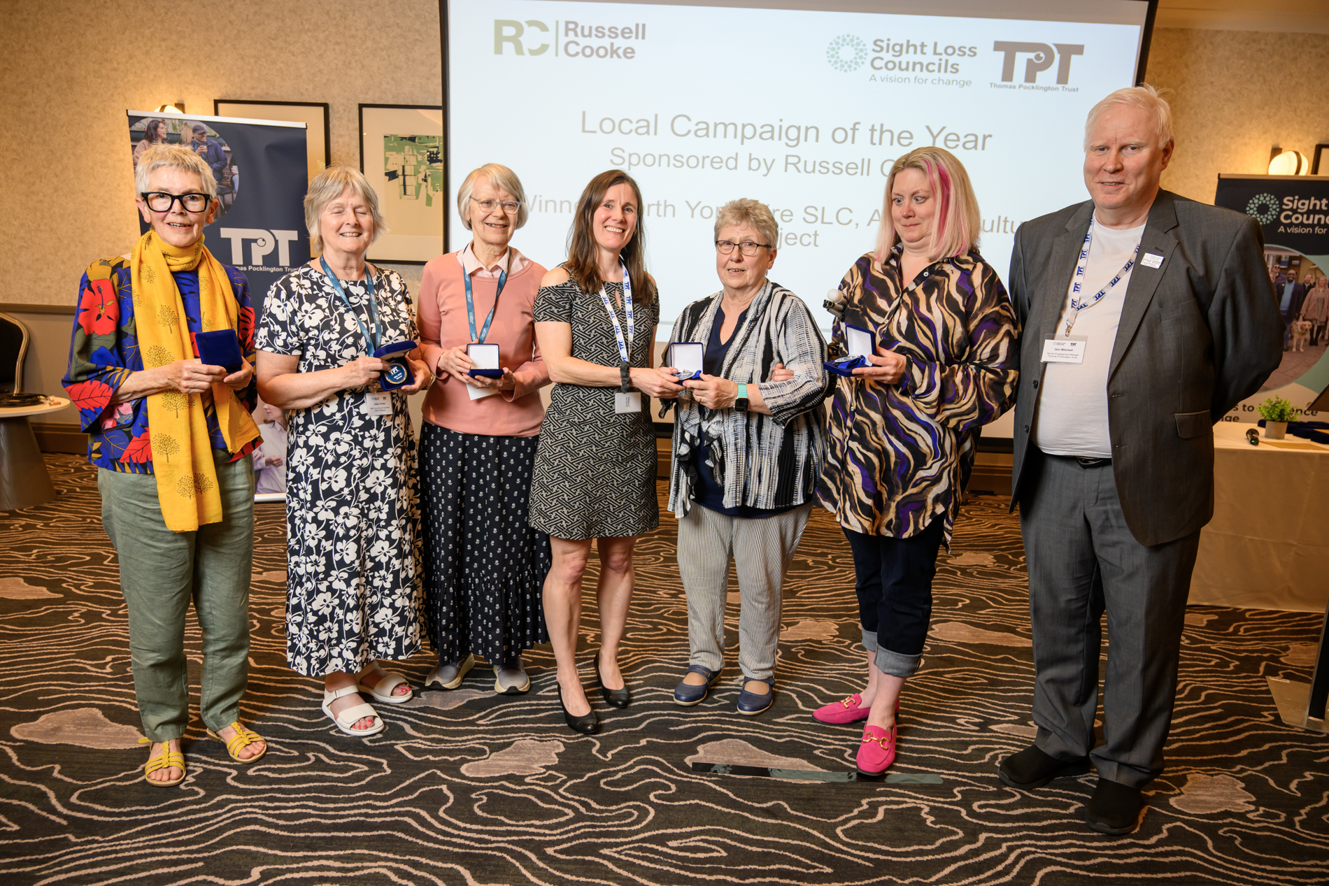 Members of North Yorkshire SLC stand together at the front of the conference room, face the camera, holding their winners medals, alongside Senior Engagement Manager Iain Mitchell. Behind the group, a large screen displays the text: 'Local Campaign of the Year - sponsored by Russell Cooke.