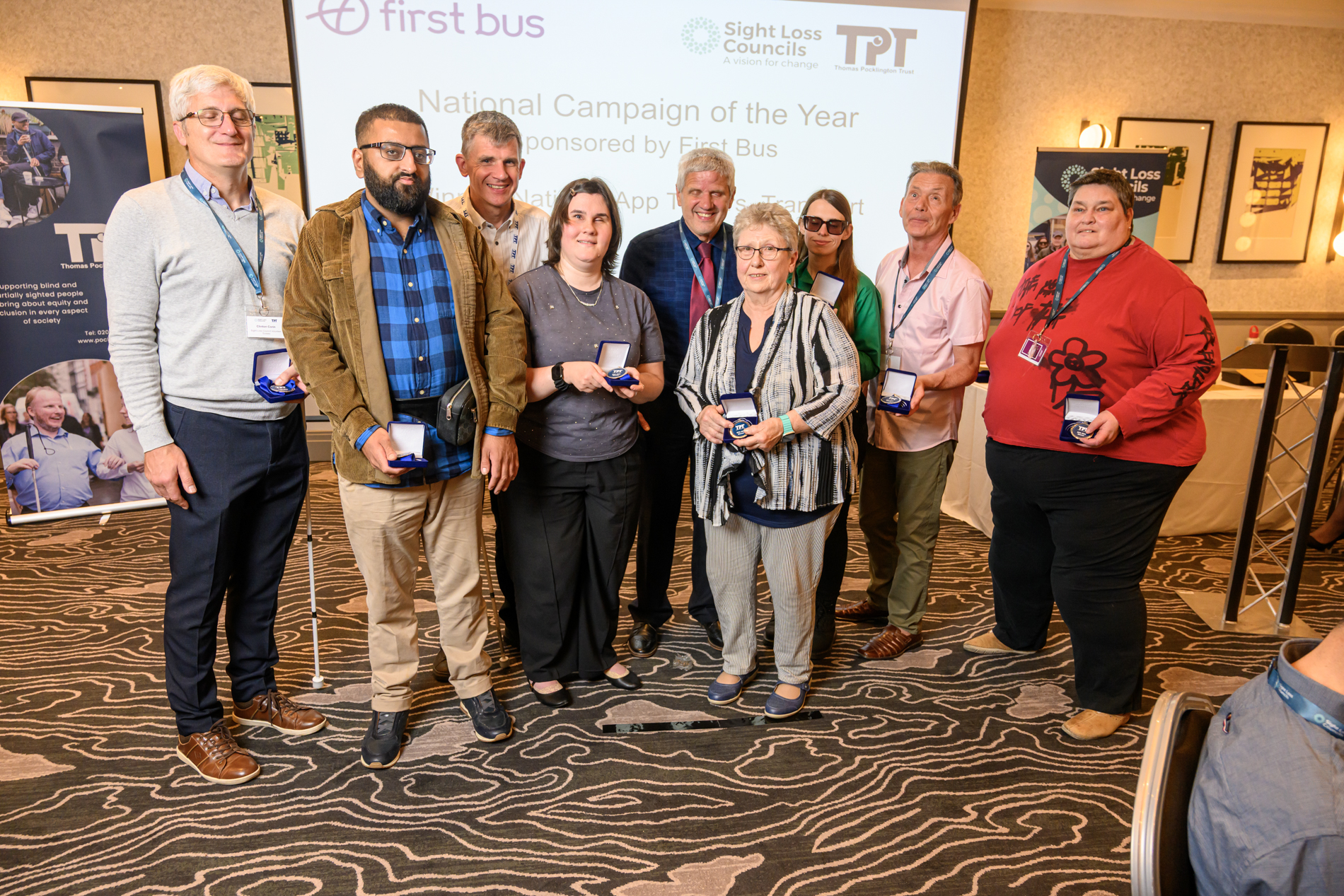 Nine members of the National App Testers group, and winners of National Campaign of the Year, stand together at the front of the Rodney Powell Awards ceremony. Behind them, a large screen displays the text “National Campaign of the Year – Sponsored by First Bus”. Members of the group each hold a medal box and is smiling towards the camera.