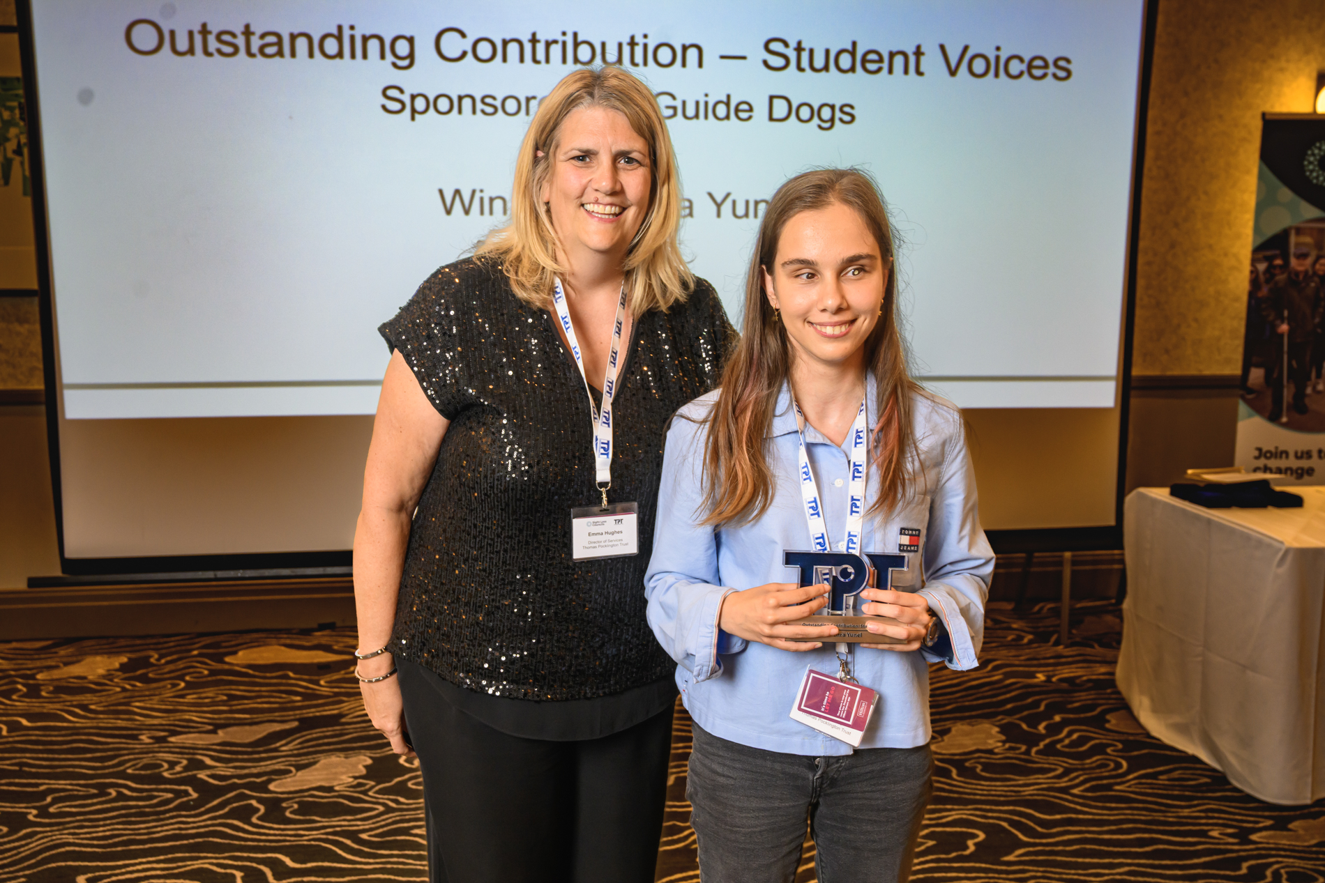 Zehra, Student Voices volunteer, stands with Director of Services, Emma Huges at the 2026 Rodney Powell Awards. Zehra is holding a TPT shaped trophy as the winner of the Outstanding Contribution to Studen Voices award. A screen behind them displays the text “Outstanding Contribution – Student Voices,” sponsored by Guide Dogs, and identifies the winner as Zehra Yunel.