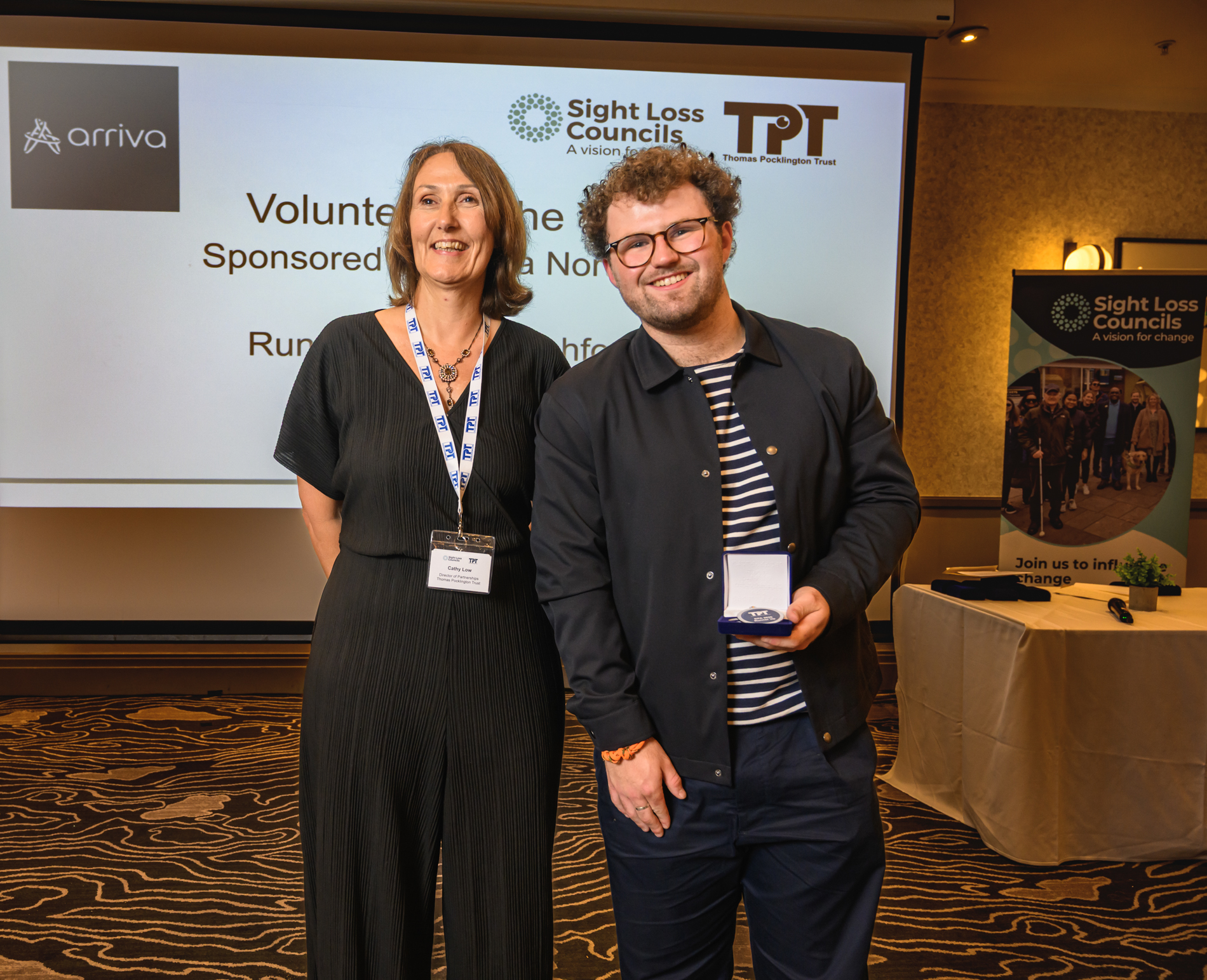 Tom Ashworth, a Young Voices volunteer and runner-up in the Volunteer of the Year category, stands with Cathy Lowe, Director of Services, at the Rodney Powell Awards. Tom holds an open presentation case containing a medal while posing in front of a large presentation screen displaying the Volunteer of the Year award category, sponsored by Arriva. Sight Loss Councils and Thomas Pocklington Trust logos are visible on the screen, with a Sight Loss Councils banner and awards table in the background.