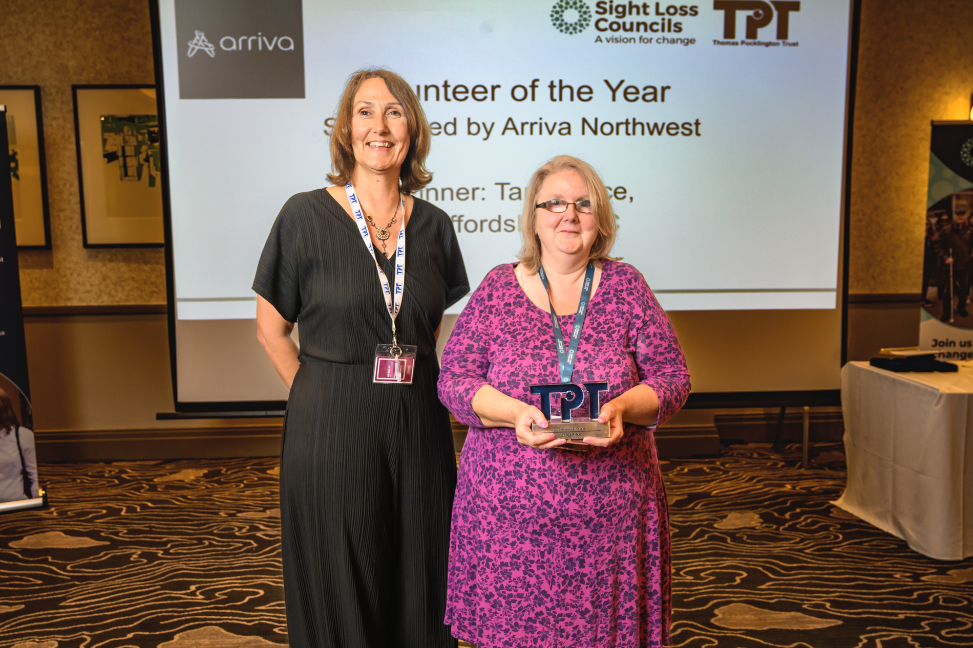 Tanja, Staffordshire SLC member and winner of the Volunteer of the Year award poses with Cathy Lowe, Director of Services, at the Rodney Powell Awards. The award recipient holds a TPT-shaped trophy while standing in front of a large presentation screen displaying the Volunteer of the Year category, sponsored by Arriva North West.